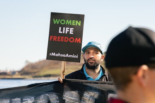 A man holding a sign that reads 'WOMEN LIFE FREEDOM #MahsaAmini'. He is wearing a blue cap and appears to be part of a protest or rally. There is another person partially visible in the foreground, and a body of water with a grassy hill in the background.