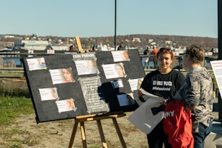A protest scene with a display board featuring profiles of individuals labeled as being connected to Evin Prison. Two people, one wearing a 'Be Our Voice' t-shirt, engage in conversation beside the board. They are outdoors in a park-like setting with a body of water and urban backdrop.