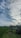A collection of abandoned vehicles is scattered in a lush green field under a sky filled with cloud patterns. The grass is tall and vibrant, surrounded by trees.
