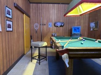 A vintage-style game room with wood-paneled walls and various framed photos. A pool table with colored balls and a cue is centered under a large ceiling light with a yellowish glow. A beige stool is near the table, and a television displaying two people is against the back wall along with decorative items like a fish mount and a landscape painting.