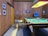 A vintage-style game room with wood-paneled walls and various framed photos. A pool table with colored balls and a cue is centered under a large ceiling light with a yellowish glow. A beige stool is near the table, and a television displaying two people is against the back wall along with decorative items like a fish mount and a landscape painting.