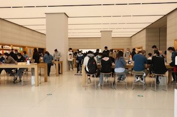 A bustling interior of a modern retail store with people seated and standing around wooden tables, interacting with electronic devices. The clean, minimalist design features a high ceiling with rectangular lighting panels. Shelves along the walls display various products and accessories.