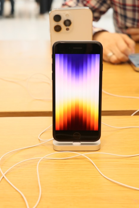 A smartphone is displayed on a wooden table, supported by a stand. The screen shows a pattern of vertical stripes in gradient colors ranging from yellow to purple. In the background, another larger smartphone is visible, with a focus on its camera lenses. A person's hand is partially visible, interacting with a device.