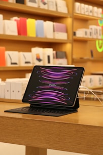 A sleek iPad Pro is displayed on a wooden table with its screen showing a dynamic pink and purple pattern. The tablet is accompanied by a keyboard case and connected to a power source via a white cable. In the background, shelves are lined with colorful product boxes and accessories, suggesting a modern retail setting.