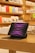 A sleek iPad Pro is displayed on a wooden table with its screen showing a dynamic pink and purple pattern. The tablet is accompanied by a keyboard case and connected to a power source via a white cable. In the background, shelves are lined with colorful product boxes and accessories, suggesting a modern retail setting.