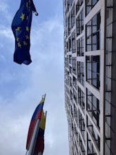 A tall modern building with a grid-like facade stretches upwards into the sky. Two flags are prominently displayed, one with yellow stars on a blue background and another featuring vertical stripes of yellow, blue, and red.
