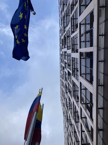 A tall modern building with a grid-like facade stretches upwards into the sky. Two flags are prominently displayed, one with yellow stars on a blue background and another featuring vertical stripes of yellow, blue, and red.