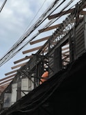 An under-construction or partially dismantled wooden building structure with exposed beams and lattice work on the roof. Visible are windows and doors along with a person wearing an orange shirt, possibly a construction worker. Power lines are prominently crossing the scene.