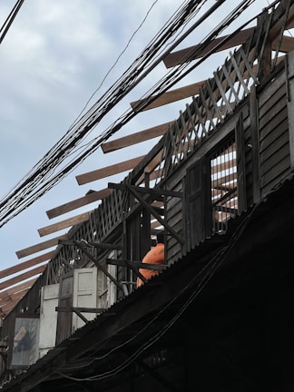 An under-construction or partially dismantled wooden building structure with exposed beams and lattice work on the roof. Visible are windows and doors along with a person wearing an orange shirt, possibly a construction worker. Power lines are prominently crossing the scene.