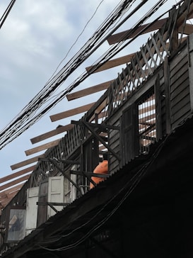 An under-construction or partially dismantled wooden building structure with exposed beams and lattice work on the roof. Visible are windows and doors along with a person wearing an orange shirt, possibly a construction worker. Power lines are prominently crossing the scene.