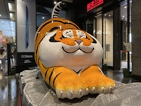 A large, cartoon-style tiger sculpture with a playful and cheerful expression sits on a textured stone platform in an indoor setting. The tiger is predominantly orange with black stripes and white accents on its face and belly. Surrounding the sculpture are commercial elements, such as a store entrance and advertising displays.