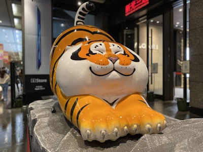 A large, cartoon-style tiger sculpture with a playful and cheerful expression sits on a textured stone platform in an indoor setting. The tiger is predominantly orange with black stripes and white accents on its face and belly. Surrounding the sculpture are commercial elements, such as a store entrance and advertising displays.