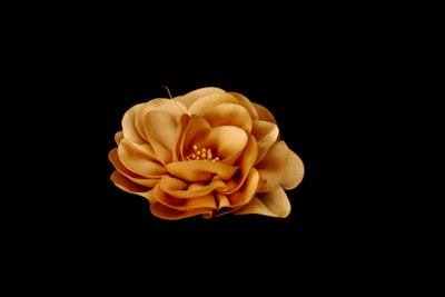 A delicate, velvet-like fabric flower with multiple layered petals in shades of soft ochre and tan, set against a black background. The central part of the flower features small, rounded, gold pearl-like beads.
