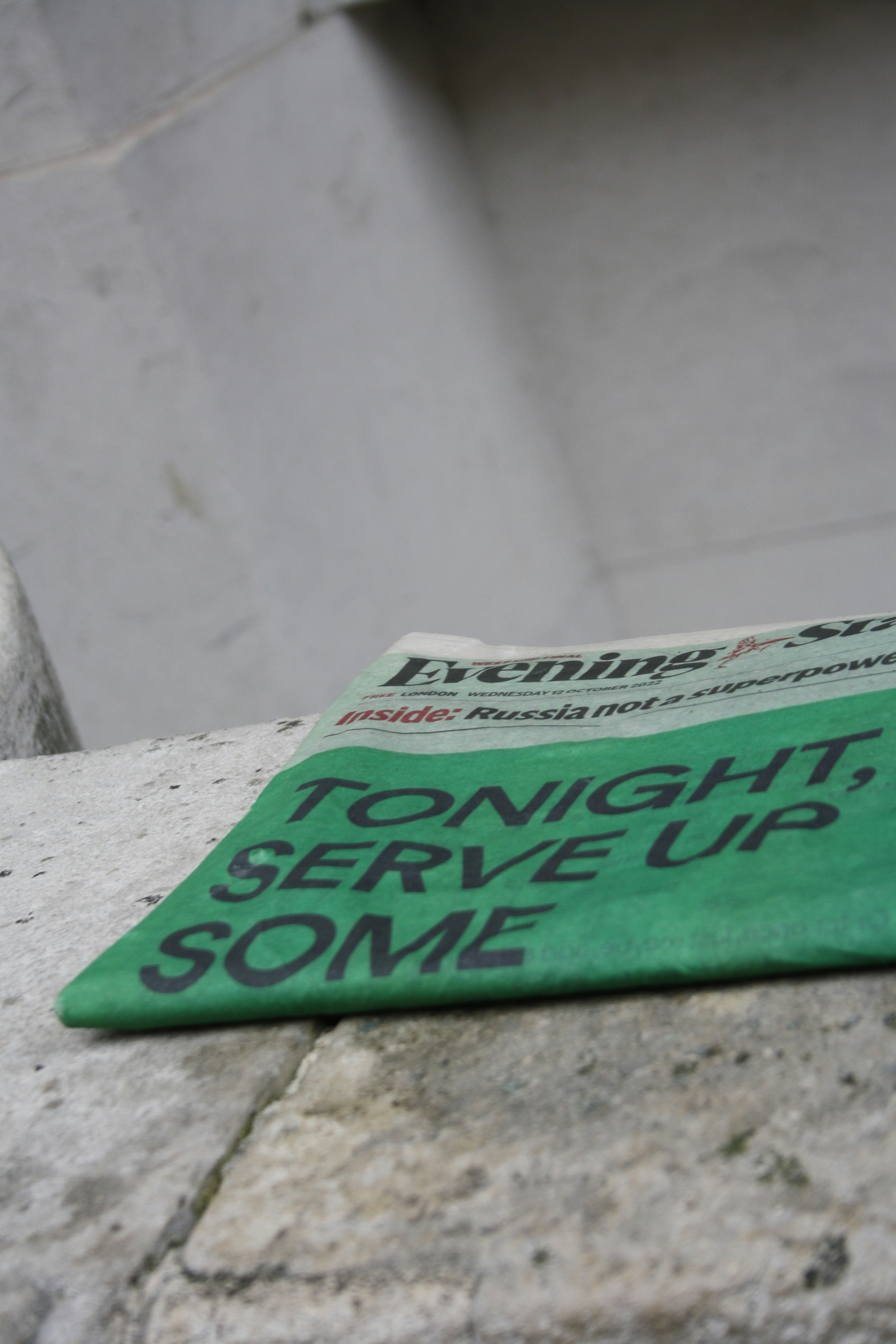 A green newspaper with bold black text suggesting an upcoming event lies angled on a rough stone surface. The background is a plain, light-colored wall, giving a stark contrast to the vibrant hue of the newspaper.