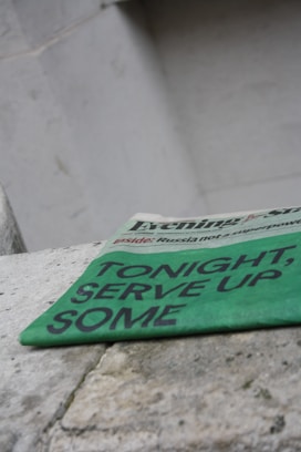 A green newspaper with bold black text suggesting an upcoming event lies angled on a rough stone surface. The background is a plain, light-colored wall, giving a stark contrast to the vibrant hue of the newspaper.