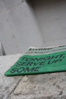 A green newspaper with bold black text suggesting an upcoming event lies angled on a rough stone surface. The background is a plain, light-colored wall, giving a stark contrast to the vibrant hue of the newspaper.