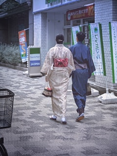 Two people are walking down a street wearing traditional Japanese clothing, with a convenience store in the background. They are carrying small bags, and flags with Japanese writing line the street.