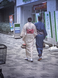 Two people are walking down a street wearing traditional Japanese clothing, with a convenience store in the background. They are carrying small bags, and flags with Japanese writing line the street.