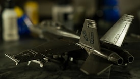 A detailed model airplane is positioned on a dark, reflective surface. The background is blurred, showing various bottles with blue, yellow, and white colors, adding a subtle depth of field effect to the image. The model airplane is metallic with intricate details and markings on its wings and tail.