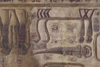 The image depicts an ancient Egyptian hieroglyphic scene featuring figures with humanoid bodies and animal heads, alongside snakes and abstract symbols. The artwork is inscribed on a worn, stone-like surface.