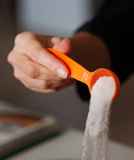A person's hand holding an orange measuring spoon filled with white granulated sugar, with some sugar spilling out.
