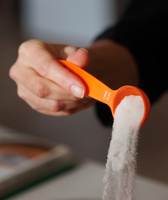 A person's hand holding an orange measuring spoon filled with white granulated sugar, with some sugar spilling out.