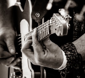 A close-up of a person playing an electric guitar. The focus is on the fingers pressing the strings and the headstock of the guitar. The person is wearing a wristband and several bracelets, suggesting a casual or artistic style. The image is in black and white, highlighting the contrast and textures of the guitar and the person's hands.