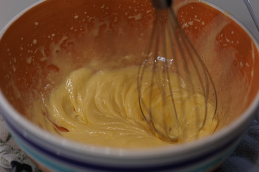 A bowl with a smooth, creamy batter being mixed with a metal whisk. The mixture has a glossy texture and is a light yellow color. The bowl is patterned with stripes on the outside and has a textured, speckled appearance on the inside.