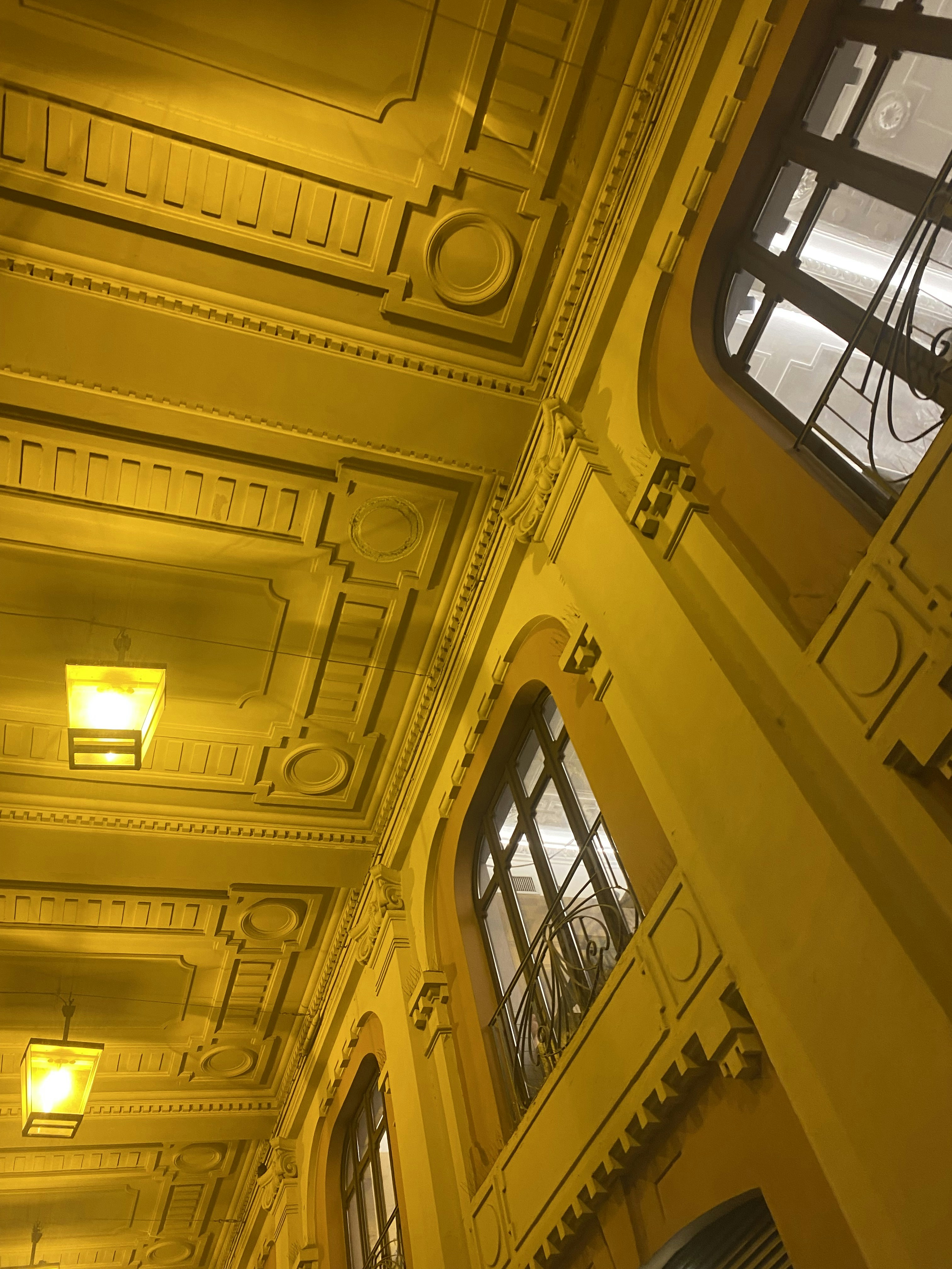 An ornate, detailed interior ceiling featuring intricate moldings and decorative designs. Three luminous rectangular light fixtures hang from the ceiling, casting a warm glow. Large windows with decorative grilles and stone arches are part of the architectural design.