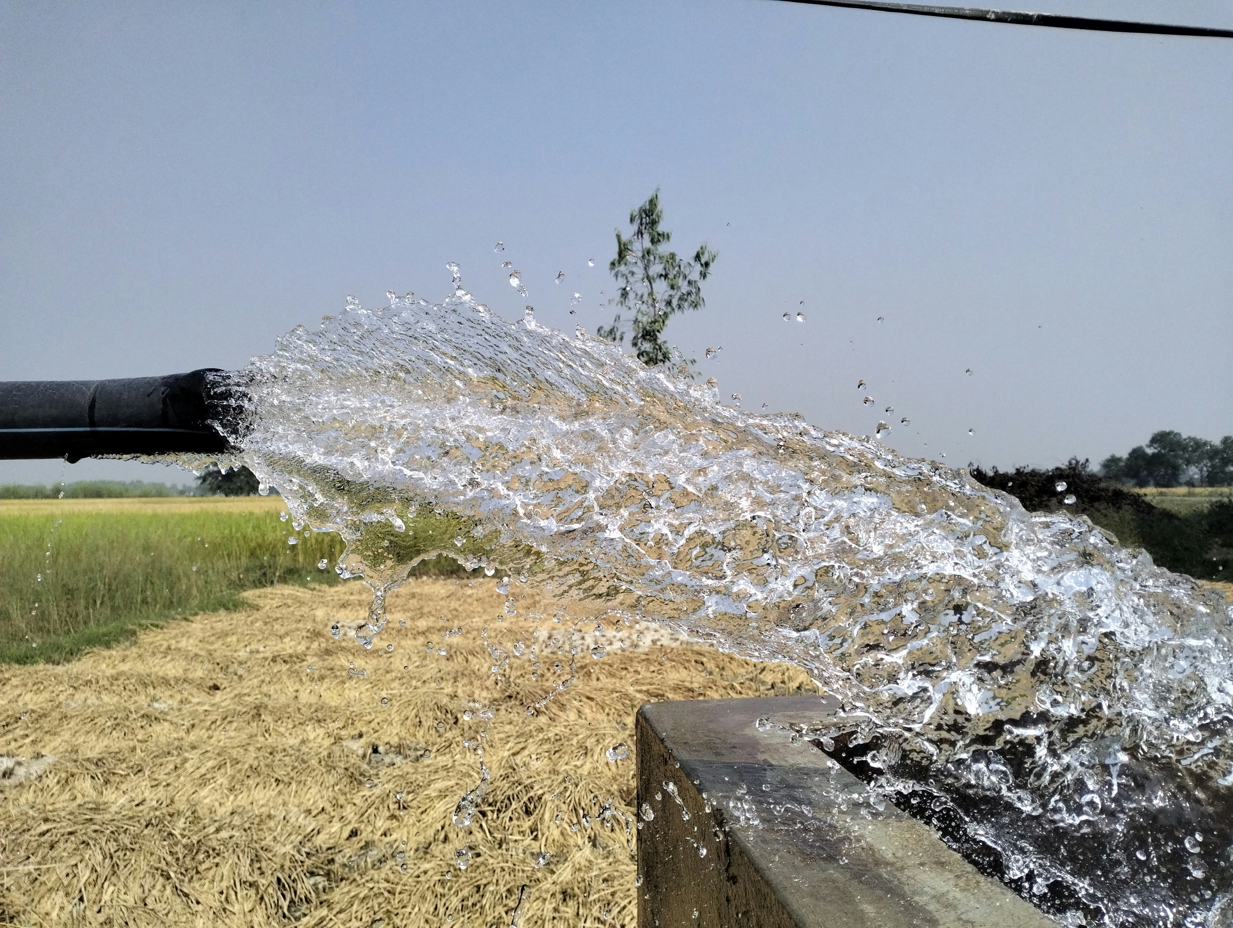 Water gushes out forcefully from a black pipe, splashing onto a concrete surface. In the background, a rural landscape with golden fields and distant trees is visible under a clear blue sky.