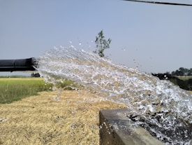 Water gushes out forcefully from a black pipe, splashing onto a concrete surface. In the background, a rural landscape with golden fields and distant trees is visible under a clear blue sky.