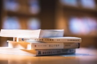 A stack of three books with Asian scripts on their spines lies on a wooden surface, under soft and warm lighting. The background is blurred, suggesting an indoor setting with shelves or more books.