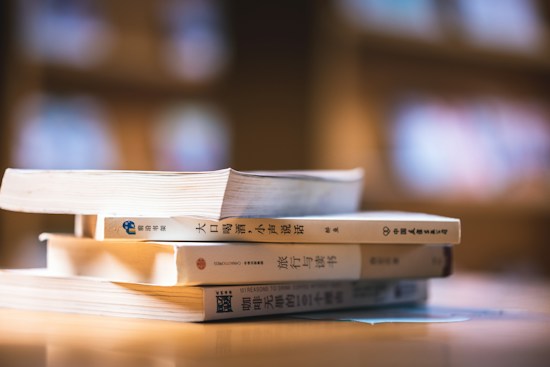 A stack of three books with Asian scripts on their spines lies on a wooden surface, under soft and warm lighting. The background is blurred, suggesting an indoor setting with shelves or more books.