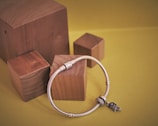 A silver bracelet adorned with charms is placed on a set of wooden blocks against a yellow background. The bracelet has a cubic metallic clasp and features an intricate bird charm.