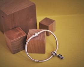 A silver bracelet adorned with charms is placed on a set of wooden blocks against a yellow background. The bracelet has a cubic metallic clasp and features an intricate bird charm.