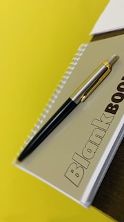 A spiral-bound notebook with the word 'Blankbook' written on its cover is placed on a yellow surface. A silver and black pen rests diagonally on top of the notebook.