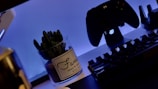 A gaming setup with a backlit keyboard in the foreground and a game controller mounted on a stand in the background. A potted cactus with a label is placed beside the keyboard. The scene is illuminated with a soft blue light, creating a relaxed atmosphere.
