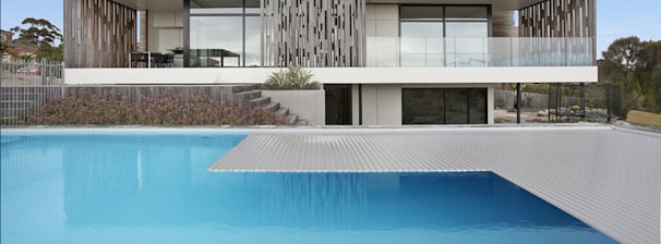 A modern house featuring a pool in the foreground. The house has a sleek design with wooden and glass elements, including a large balcony and floor-to-ceiling windows. The pool area is surrounded by landscaping with plants and a tiered garden on one side. The scene captures both the structure and natural environment.
