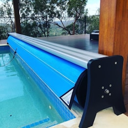 A swimming pool area featuring a blue pool cover rolled onto a metal frame at the edge of the pool. The deck is made of wooden planks, and there is a black fence with trees visible in the background. The area appears clean and well-maintained, with the cover neatly aligned along the pool's edge.