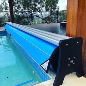 A swimming pool area featuring a blue pool cover rolled onto a metal frame at the edge of the pool. The deck is made of wooden planks, and there is a black fence with trees visible in the background. The area appears clean and well-maintained, with the cover neatly aligned along the pool's edge.