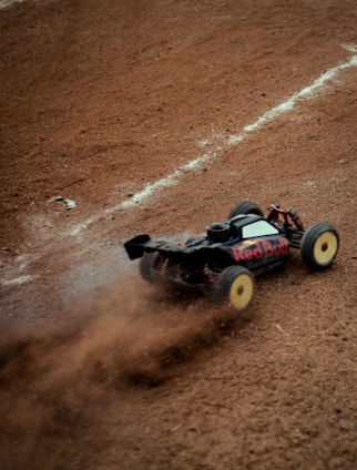 A remote-controlled car kicks up dirt as it speeds across a rough, brown dirt track. The car features prominent branding and is captured in motion, creating a dynamic and intense scene.