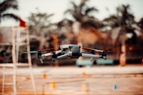A drone hovers in the air with blurred trees and objects in the background, suggesting an outdoor setting. The drone's design includes visible propellers and a camera mounted at the front, indicating its potential use for aerial photography or videography.