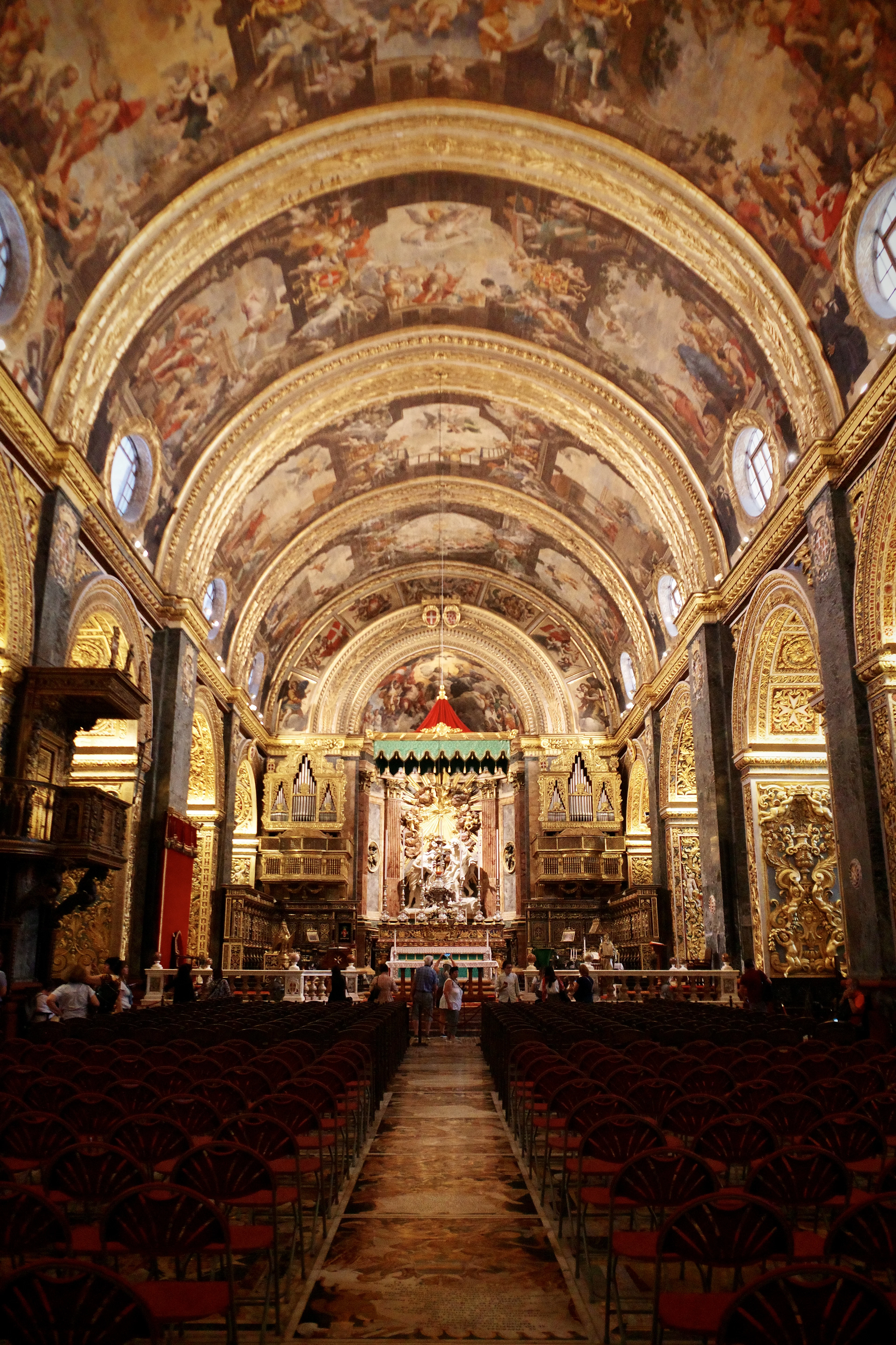 The nave of St John's Co-Cathedral in Valletta, its barrel-vaulted ceiling covered in painted frescoes and gilded carving above the marble tomb-slab floor and rows of red seating.