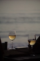Two glasses of white wine are placed on a table with a blurred background of the ocean at dusk. The scene conveys a sense of tranquility and intimacy, with soft lighting enhancing the reflective surface of the wine.