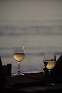 Two glasses of white wine are placed on a table with a blurred background of the ocean at dusk. The scene conveys a sense of tranquility and intimacy, with soft lighting enhancing the reflective surface of the wine.