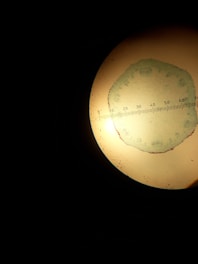 A circular magnified view through a microscope showing a greenish slide with small structures and a measuring scale overlayed across the diameter. The background is predominantly dark.