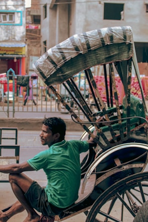 A man in a green shirt is sitting on a black rickshaw, parked on an urban street. The rickshaw has a protective cover over the seat area. The background includes concrete buildings with colorful elements, such as a red and pink cloth hanging and a yellow shop sign.