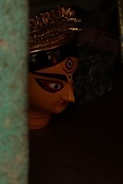A large, painted face of a traditional goddess mask with intricate eye makeup and a pointed nose. It has a golden crown with elaborate designs sitting atop the head. The image is dimly lit, adding to the mysterious and sacred atmosphere.
