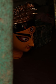 A large, painted face of a traditional goddess mask with intricate eye makeup and a pointed nose. It has a golden crown with elaborate designs sitting atop the head. The image is dimly lit, adding to the mysterious and sacred atmosphere.