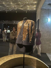 A sleek black mannequin dressed in a deep burgundy, gold-accented prom gown under soft moody lighting.